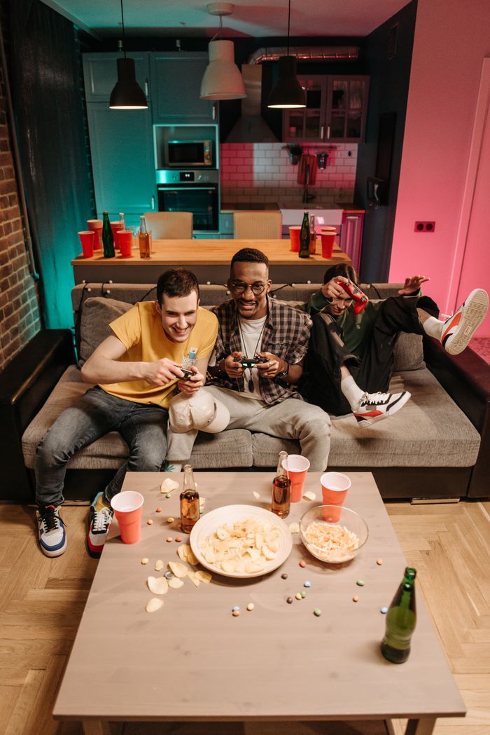 Friends enjoy video games on a couch with snacks and drinks, creating a lively living room scene.